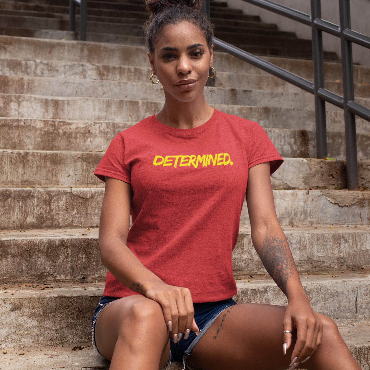 Motivational gym shirt in heather red color and yellow letters worn by a young fit woman sitting on a staircase outside the gym