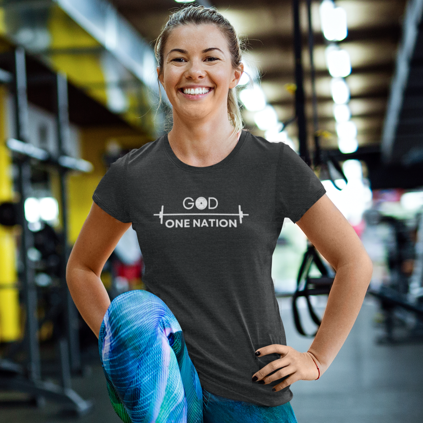 Motivational gym shirt with the words one nation and with a gym barbell graphic on a smiling woman doing a leg workout at the gym