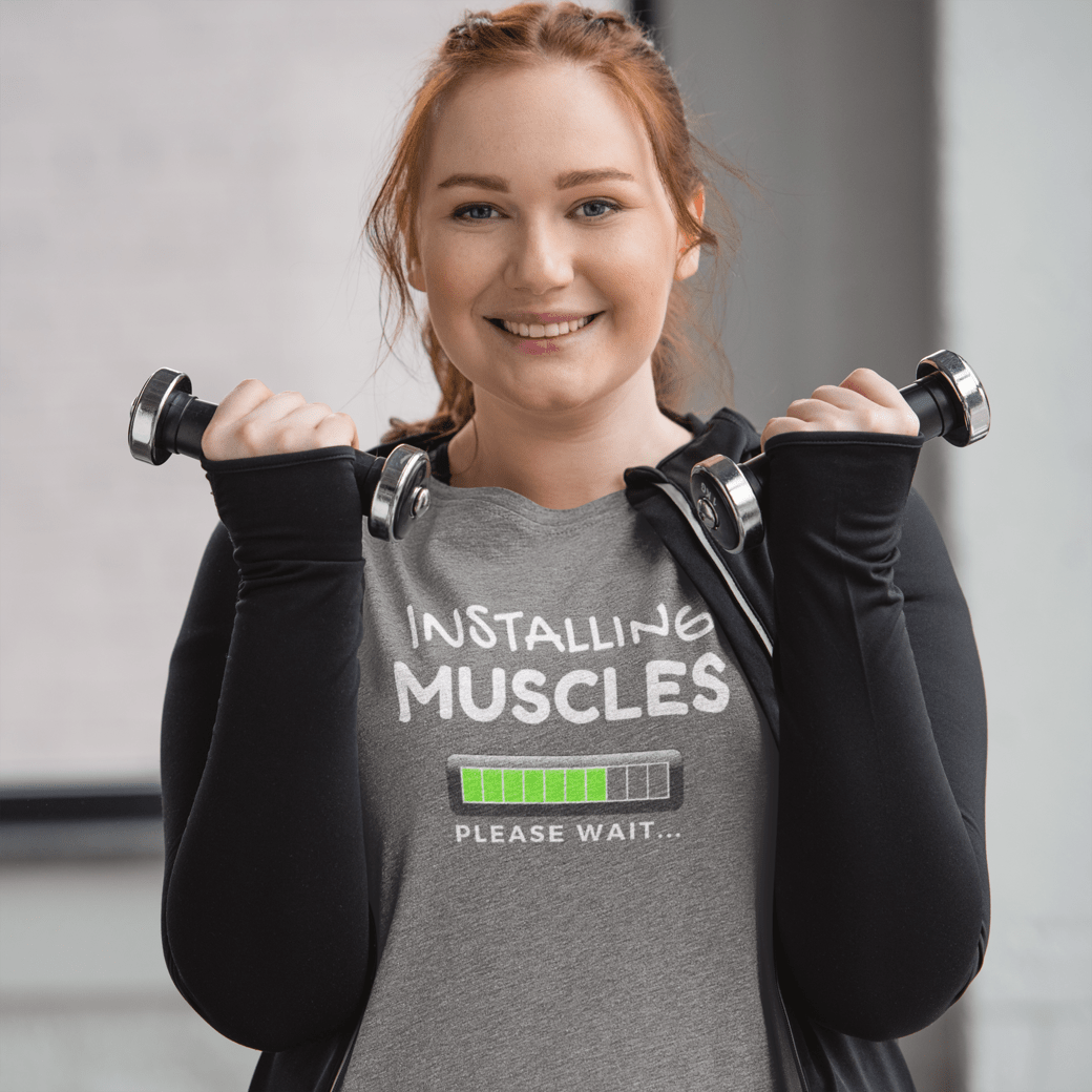 Funny gym shirt with text installing muscles please wait worn by a smiling woman lifting dumbbells